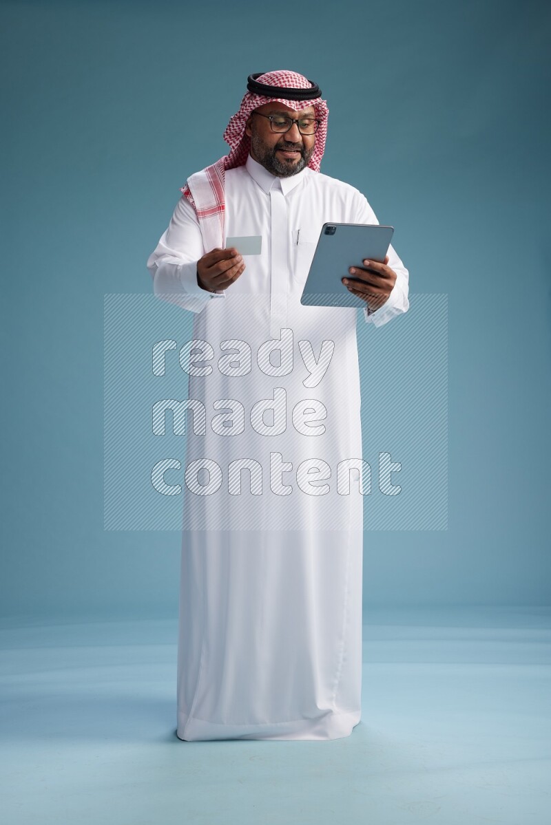 Saudi Man with shimag Standing holding ATM card while working on tablet on blue background