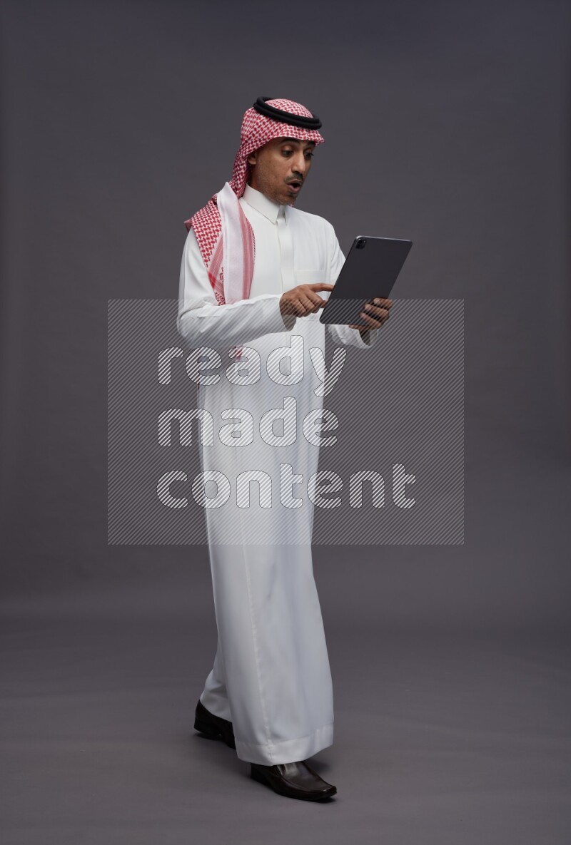 Saudi man wearing thob and shomag standing working on tablet on gray background