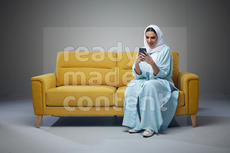 A Saudi woman wearing a light blue Abaya and white head scarf sitting on a yellow sofa and texting on her phone on a grey background