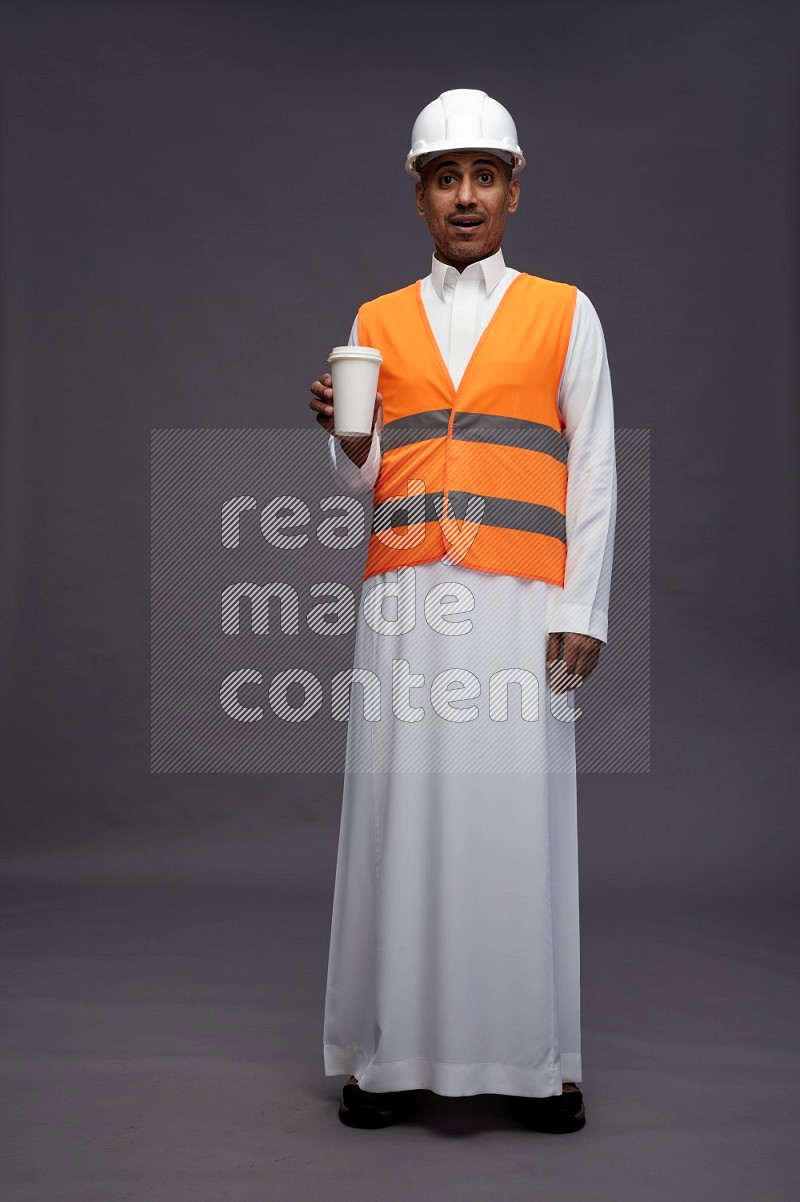 Saudi man wearing thob with engineer vest standing holding paper cup on gray background