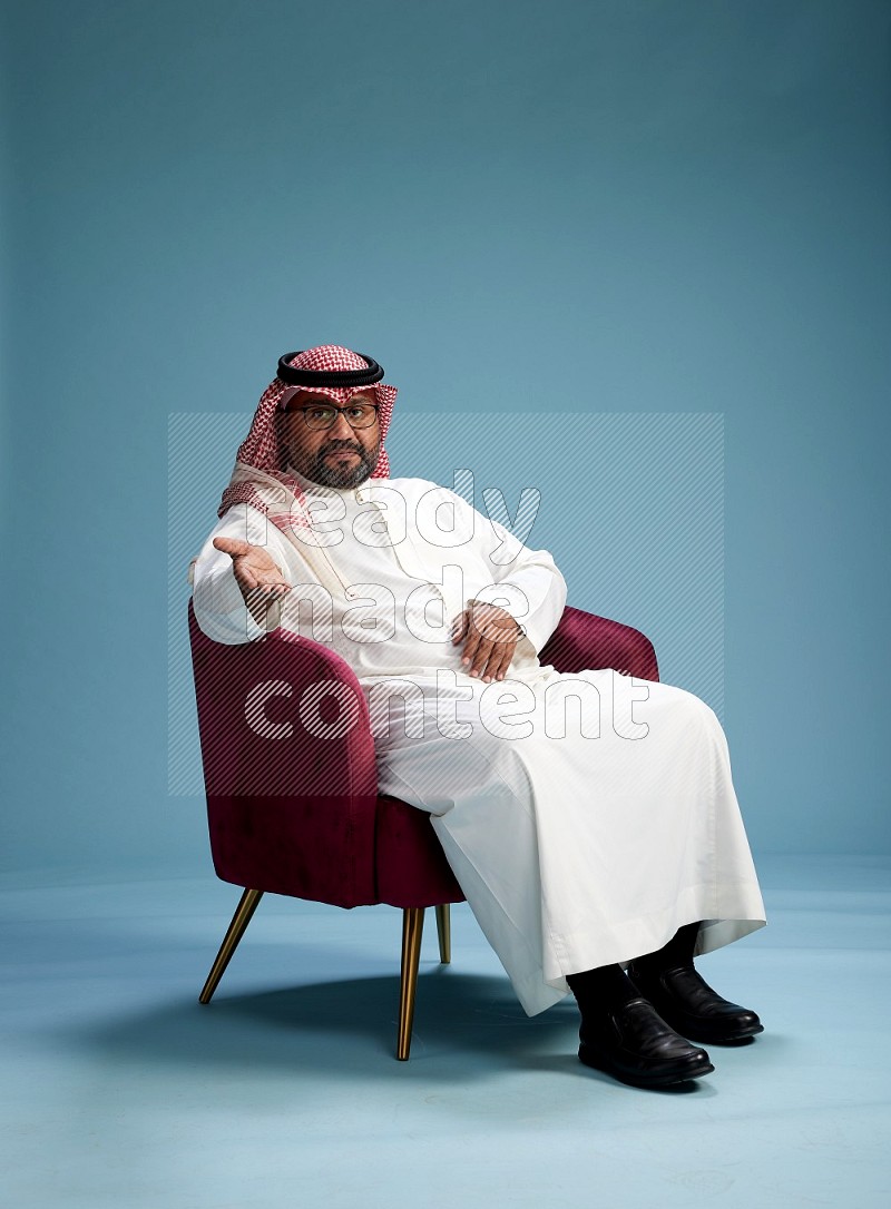 Saudi Man with shimag sitting on chair Interacting with the camera on blue background