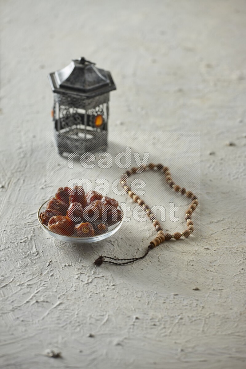 A silver lantern with different drinks, dates, nuts, prayer beads and quran on textured white background