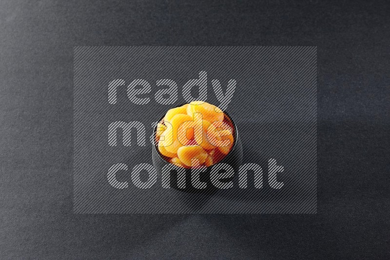 A black pottery bowl full of dried apricots on a black background in different angles