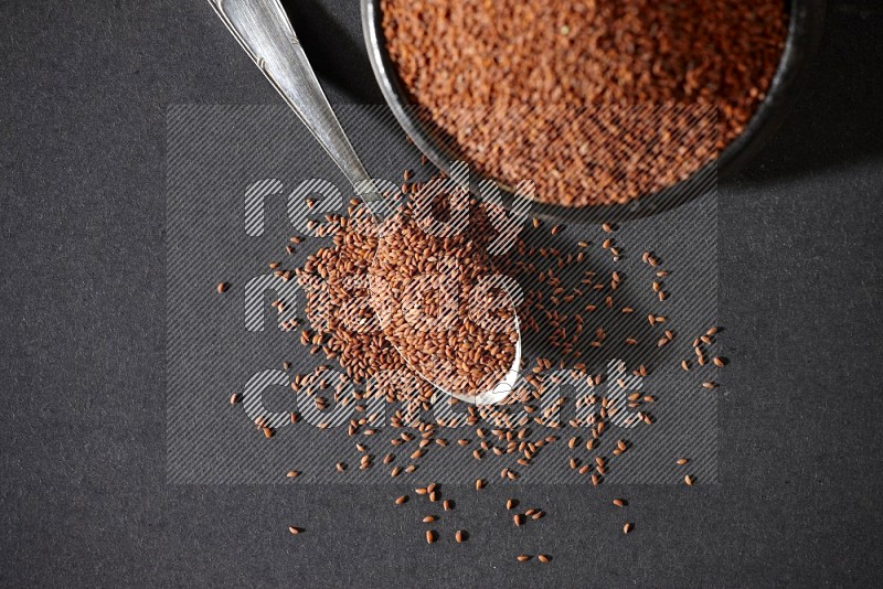 A black pottery bowl full of garden cress seeds and metal spoon full of the seeds on a black flooring