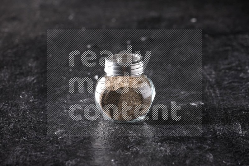 A glass spice jar full of black pepper powder on textured black flooring