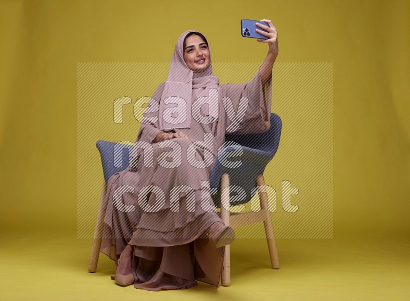 A woman sitting on a blue chair and Taking a Selfie  Setting  on a Yellow Background wearing Brown Abaya with Hijab
