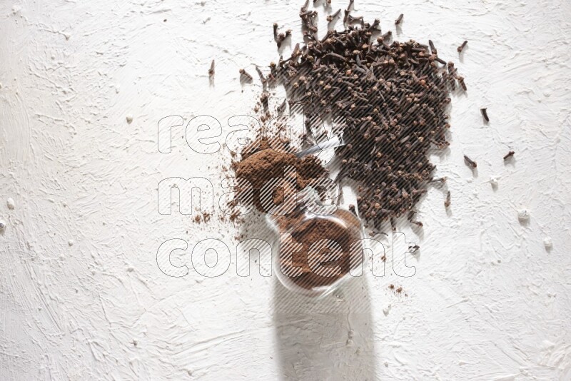 A flipped glass spice jar and a metal spoon full of cloves powder and powder came out of the jar with cloves spread on textured white flooring