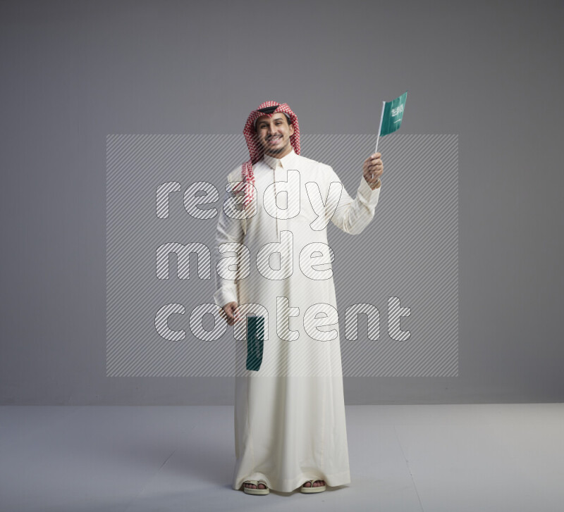 A Saudi man standing wearing thob and red shomag raising small Saudi flag on gray background