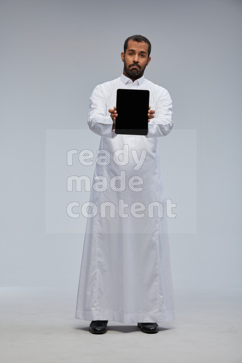 Saudi man wearing Thob standing showing tablet to camera on Gray background