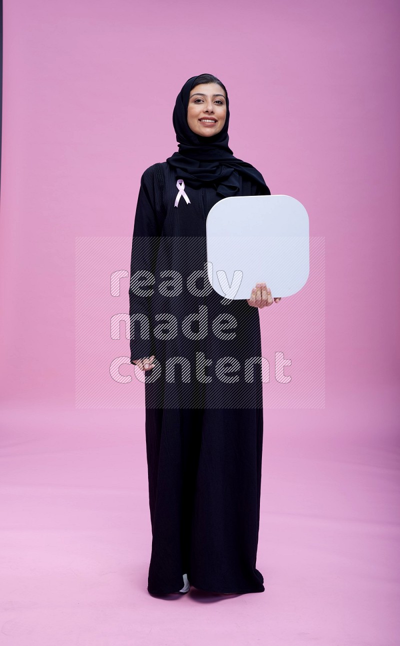 Saudi woman wearing pink ribbon on Abaya standing holding social media on pink background