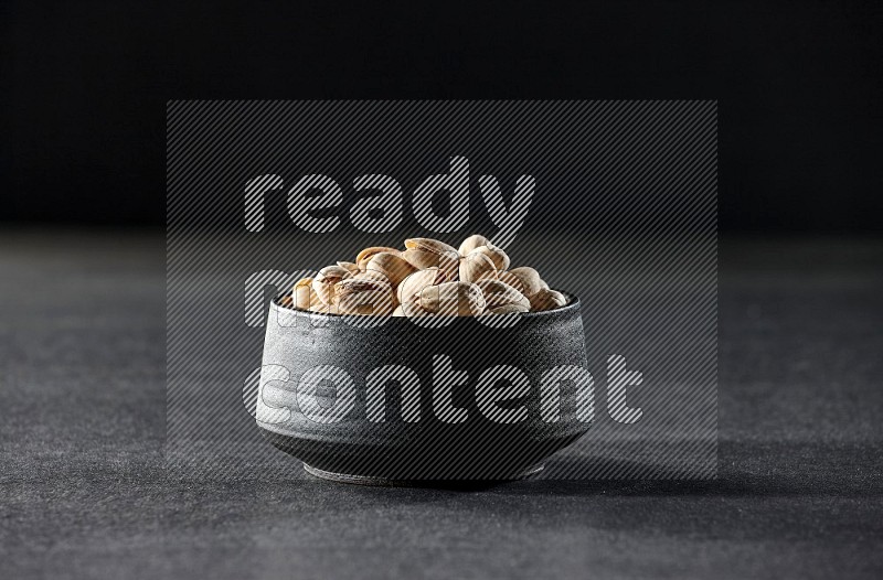 A black ceramic bowl full of pistachios on a black background in different angles