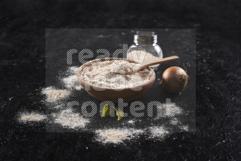 A wooden bowl full of onion powder with a glass jar beside it and fresh onion on black background