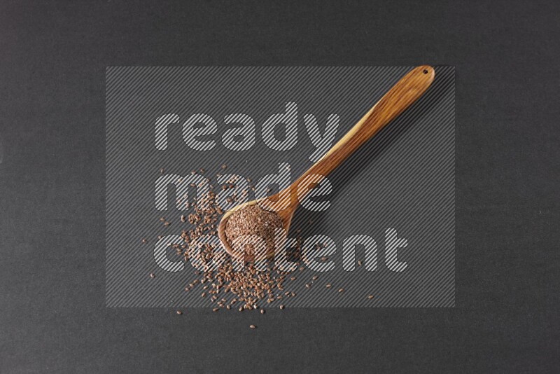 A wooden ladle full of flaxseeds and seeds spread beside it on a black flooring