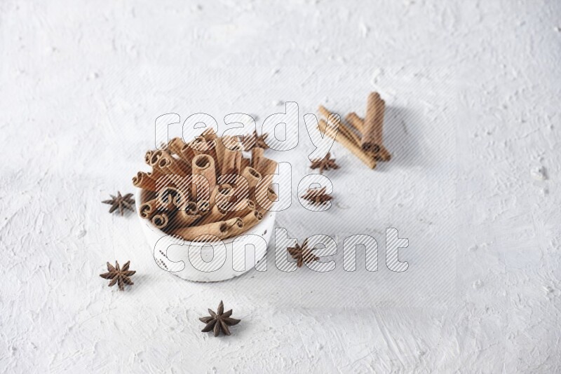 White bowl full of cinnamon sticks surrounded by star anis on a textured white background in different angles