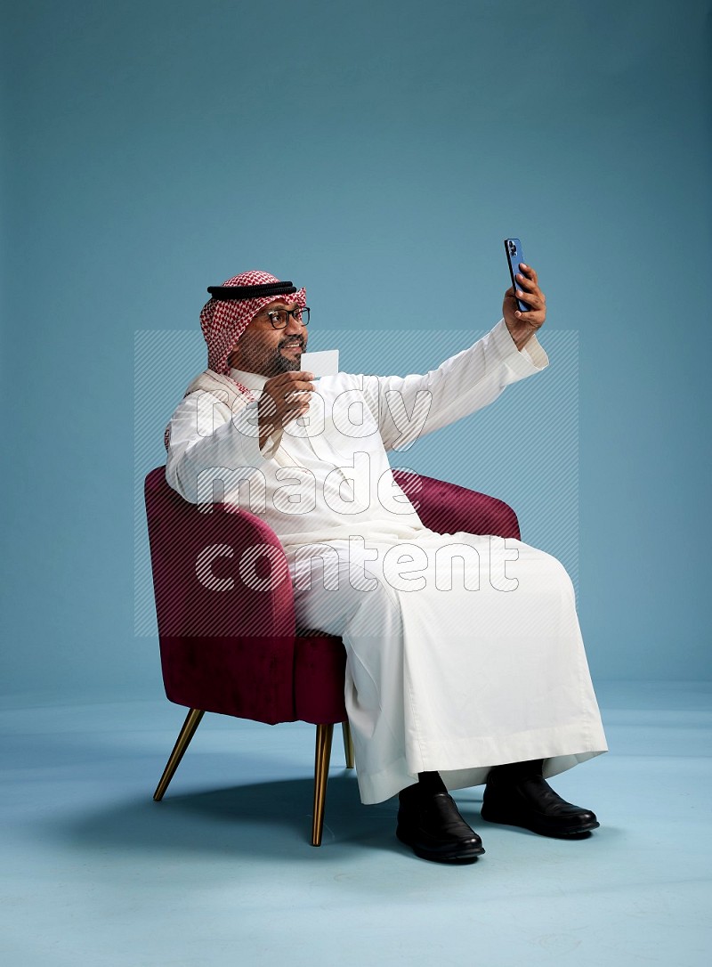 Saudi Man with shimag sitting on chair holding ATM card while talking on phone on blue background