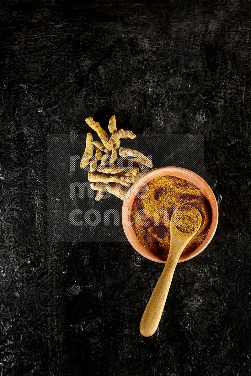 A wooden bowl and a wooden spoon full of turmeric powder with dried turmeric fingers on textured black flooring