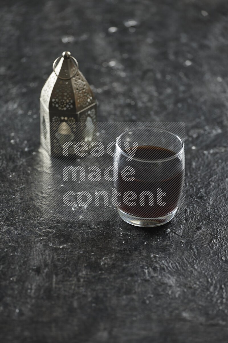 A silver lantern with different drinks, dates, nuts, prayer beads and quran on textured black background