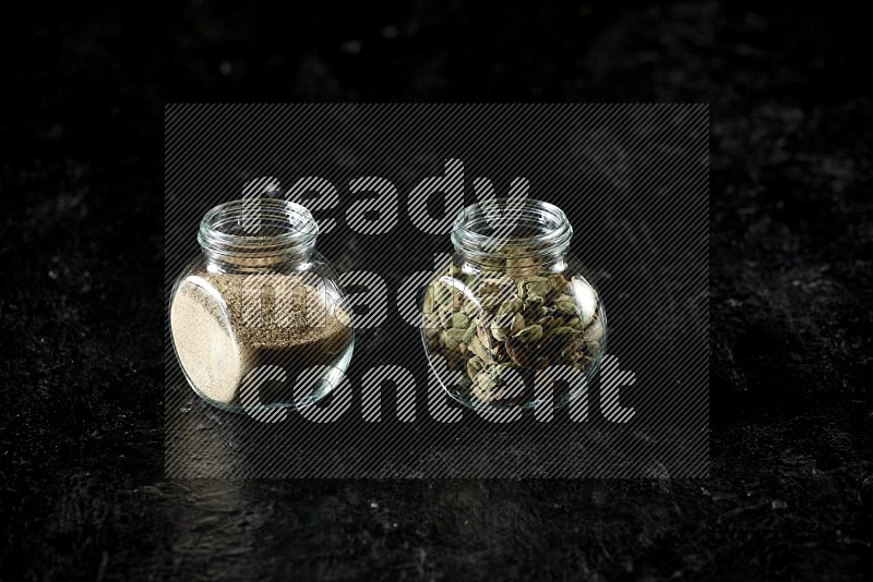 2 glass spice jars full of cardamom powder and cardamom seeds on textured black flooring