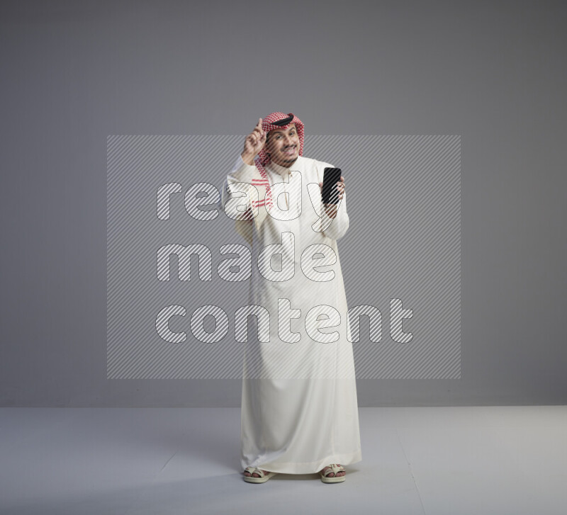 A Saudi man standing wearing thob and red shomag showing phone to camera on gray background