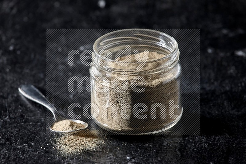 A glass jar and a metal spoon full of white pepper powder on textured black flooring