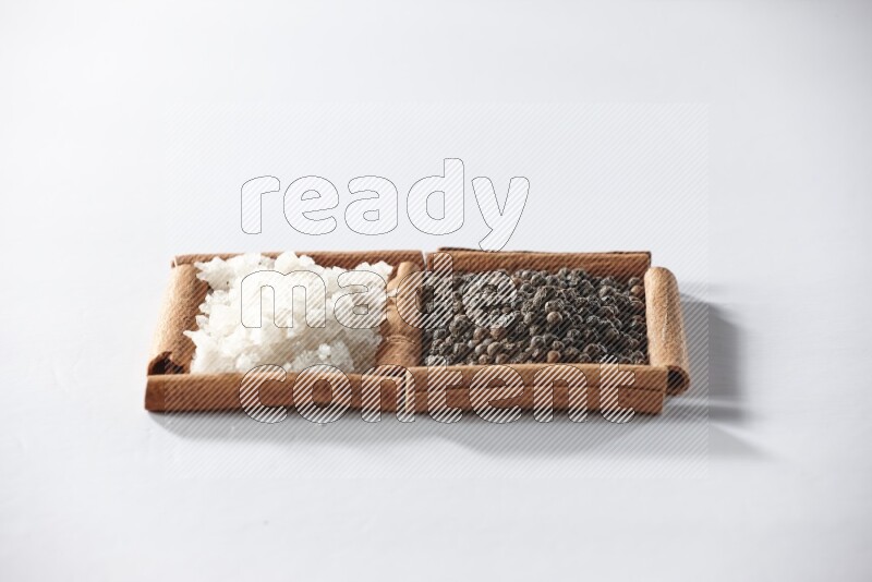 مربعان من أعواد القرفة ممتلئان بحبوب الفلفل الأسود والملح الأبيض علي خلفية بيضاء