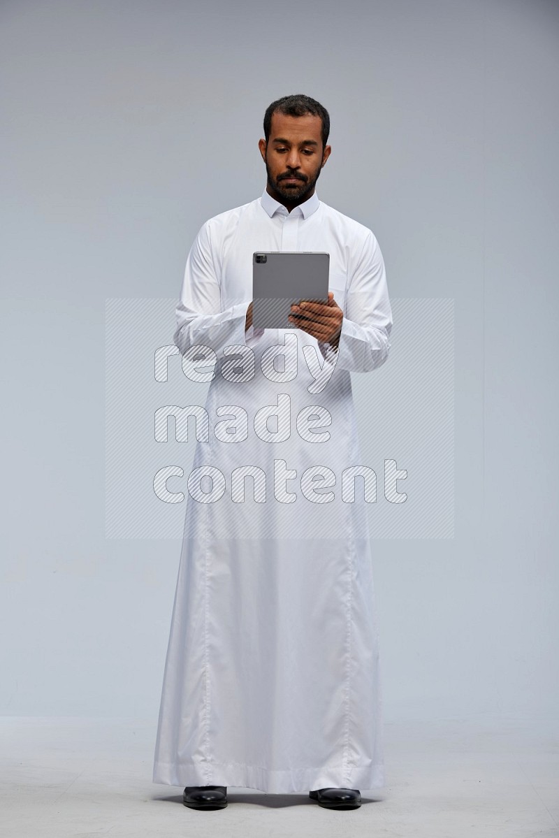 Saudi man wearing Thob standing working on tablet on Gray background