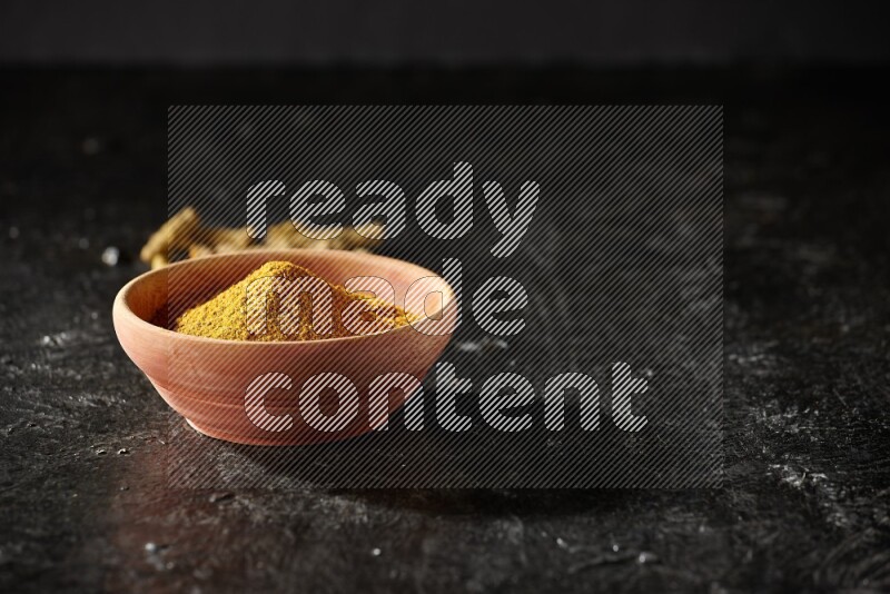 A wooden bowl full of turmeric powder with dried turmeric fingers on textured black flooring