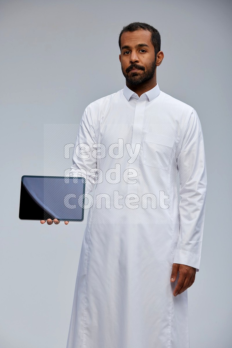 Saudi man wearing Thob standing showing tablet to camera on Gray background