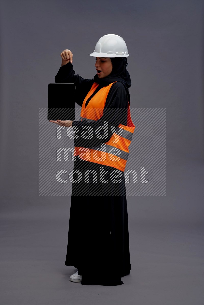 Saudi woman wearing Abaya with engineer vest standing showing tablet to camera on gray background