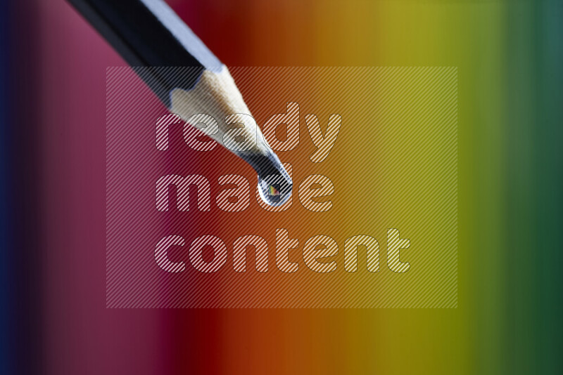 The image captures a close-up of a water droplet balanced on the sharp tip of a pencil, set against a blurred colorful background