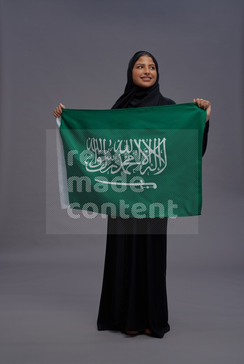 Saudi woman wearing Abaya standing holding Saudi flag on gray background