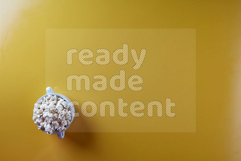 A white ceramic bowl full of popcorn on a yellow background in a top view shot