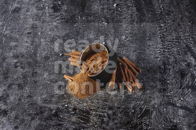 Black pottery bowl over filled with cinnamon powder and cinnamon sticks around the bowl on a textured black background