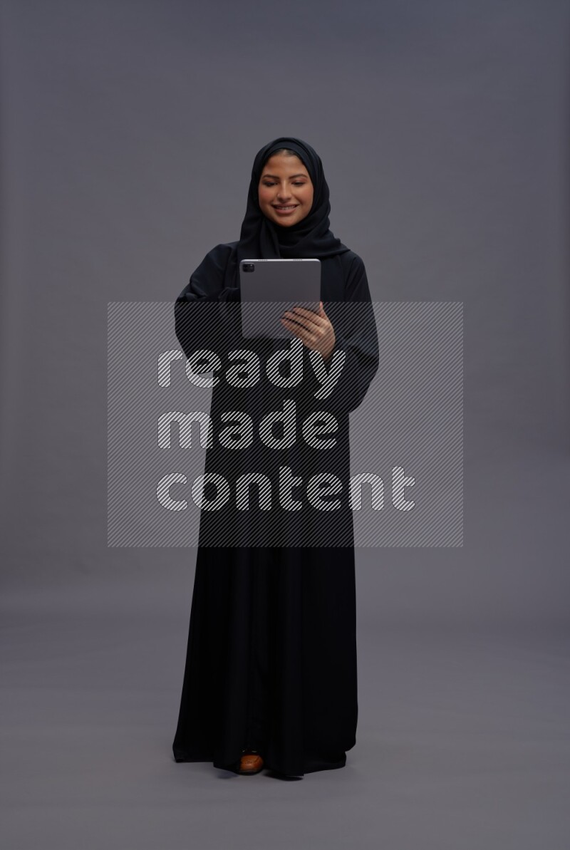 Saudi woman wearing Abaya standing working on tablet on gray background