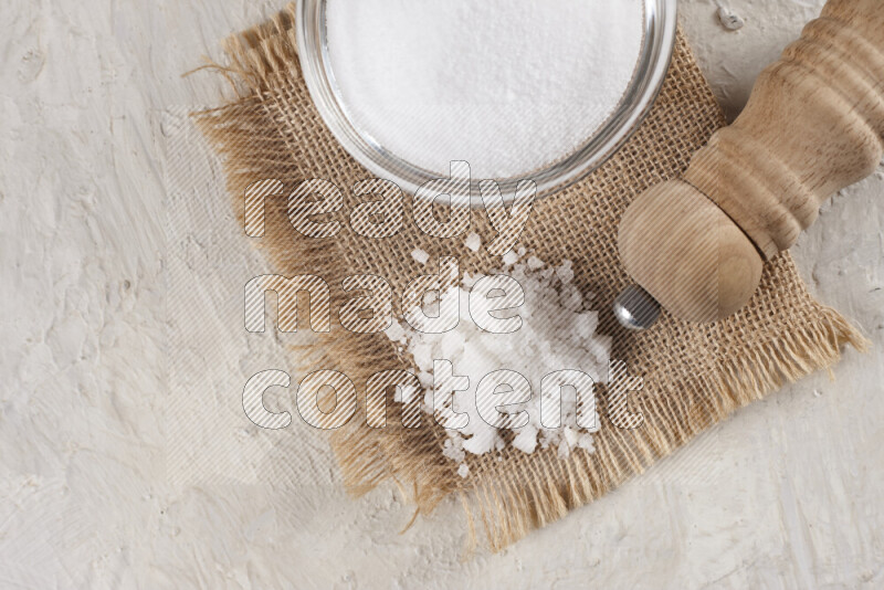 A glass bowl full of white salt with a wooden grinder on a burlap fabric all on white background