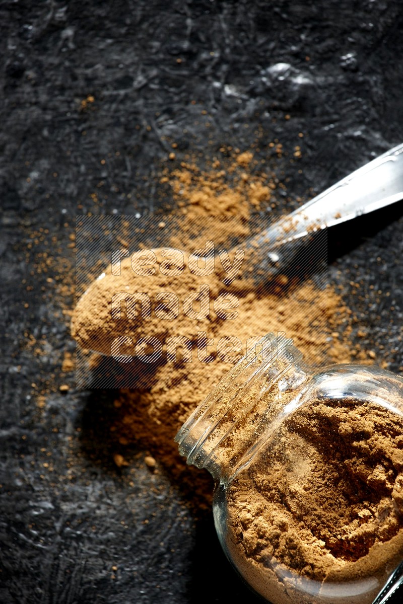 A flipped glass spice jar and a metal spoon full of allspice powder and powder spilled out of it on a textured black flooring