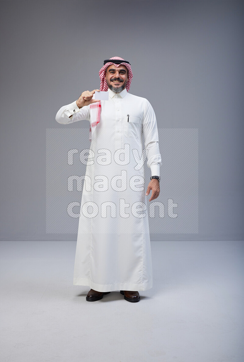 Saudi man Wearing Thob and red Shomag standing holding ATM card on Gray background