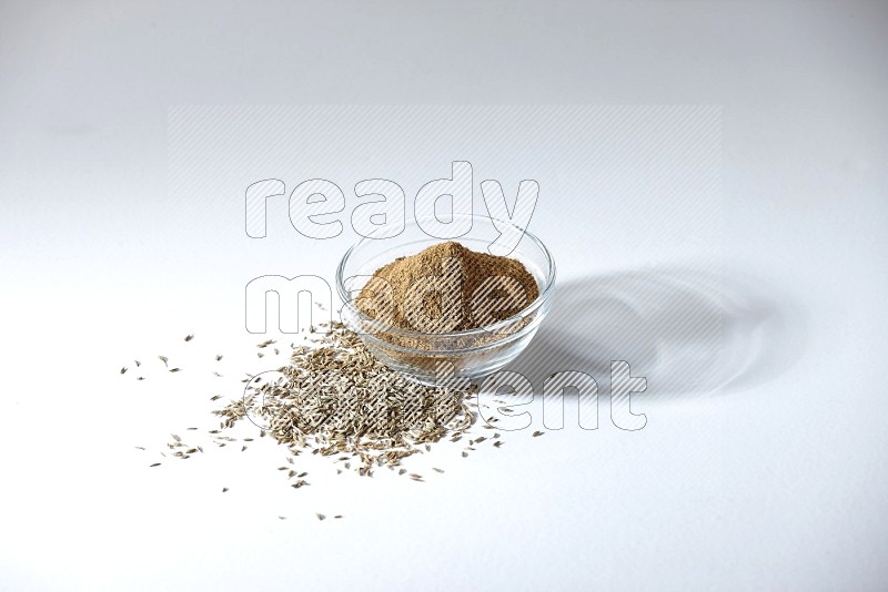 A glass bowl full of cumin powder with cumin seeds beneath it on white flooring