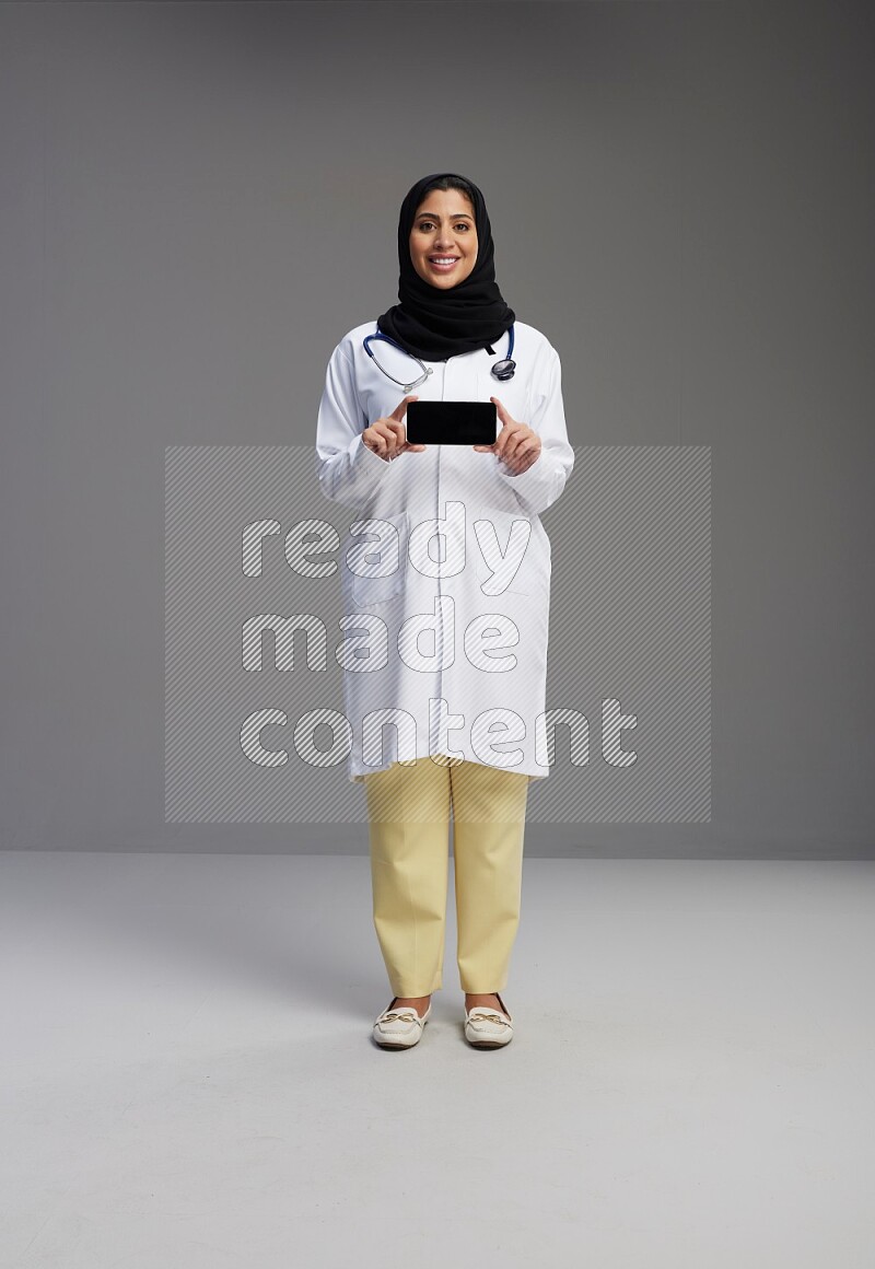 Saudi woman wearing lab coat with stethoscope standing showing phone to camera with sign in the back on Gray background