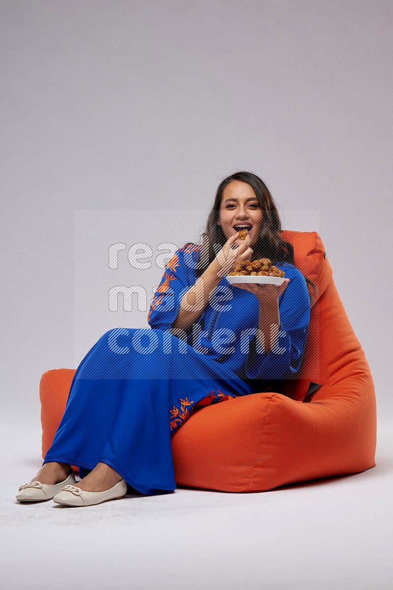 A Woman sitting on an orange beanbag wearing Jalabeya holding a plate of dates