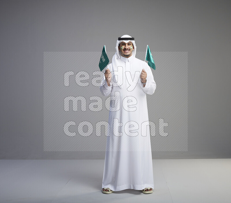 A Saudi man standing wearing thob and white shomag with face painting raising small saudi flag on gray background