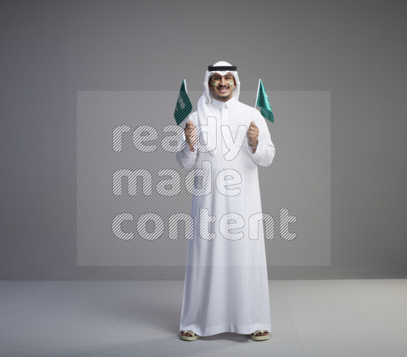 A Saudi man standing wearing thob and white shomag with face painting raising small saudi flag on gray background