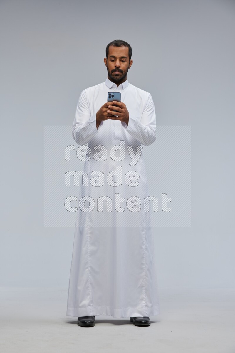 Saudi man wearing Thob standing texting on phone on Gray background