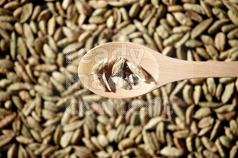 Cardamom seeds in a wooden spoon above cardamom seeds background on black flooring