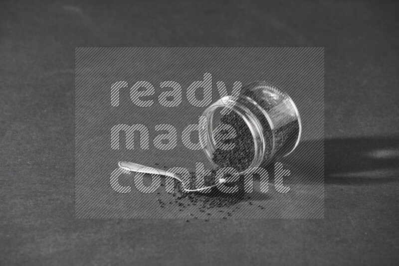 A flipped glass jar full of black seeds and seeds spread out with a metal spoon full of the seeds on a black flooring