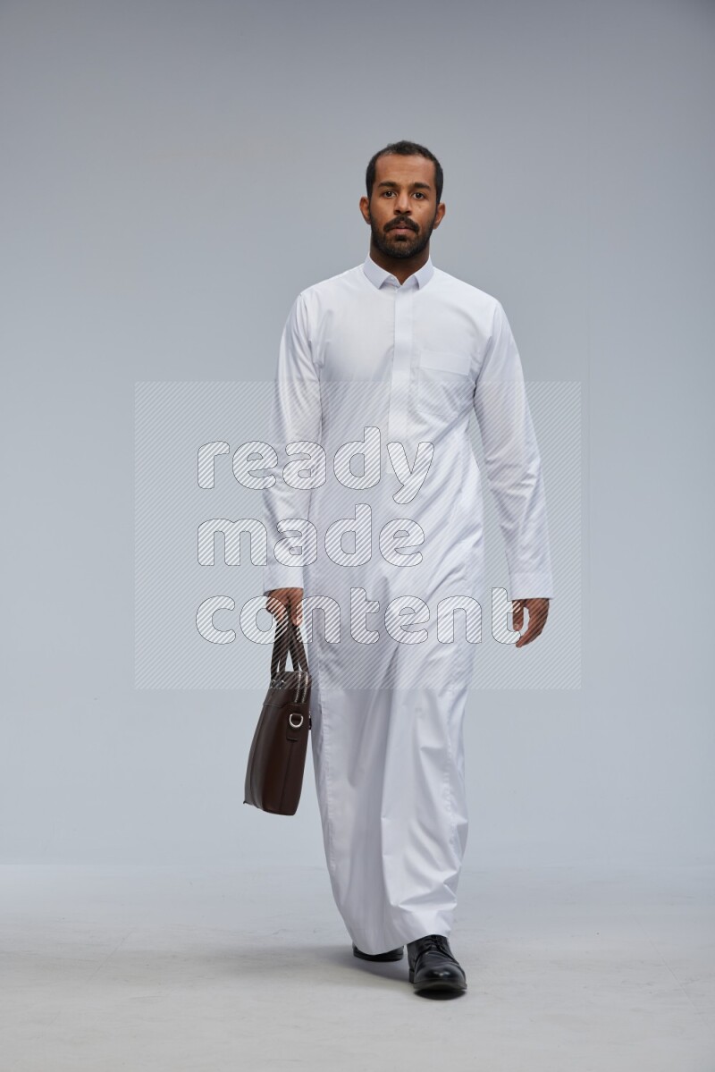 Saudi man wearing thob standing holding suitcase on gray background