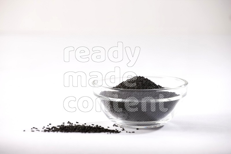 A glass bowl full of black seeds and some more seeds spread next to it on a textured white flooring