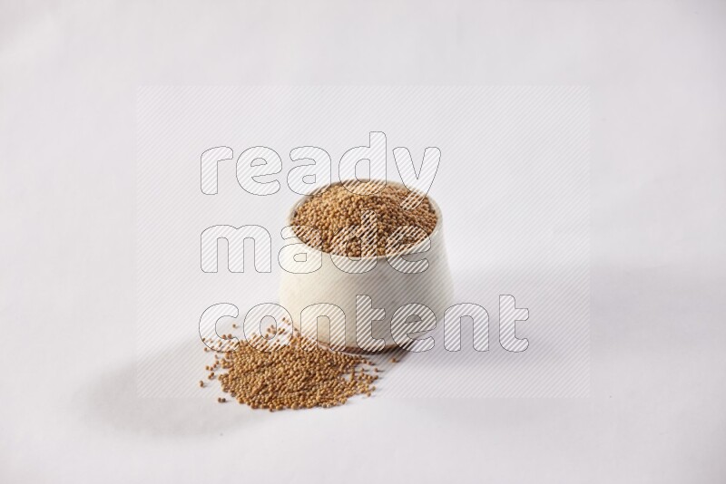 A beige pottery bowl full of mustard seeds and more seeds spread on a white flooring