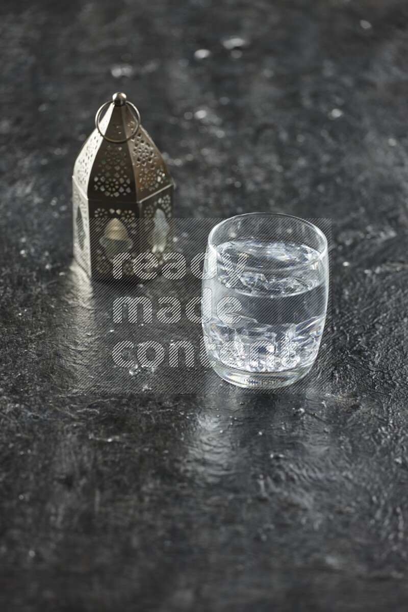 A silver lantern with different drinks, dates, nuts, prayer beads and quran on textured black background
