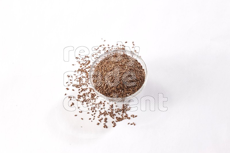 A glass bowl full of flax seeds surrounded by flax seeds on a white flooring
